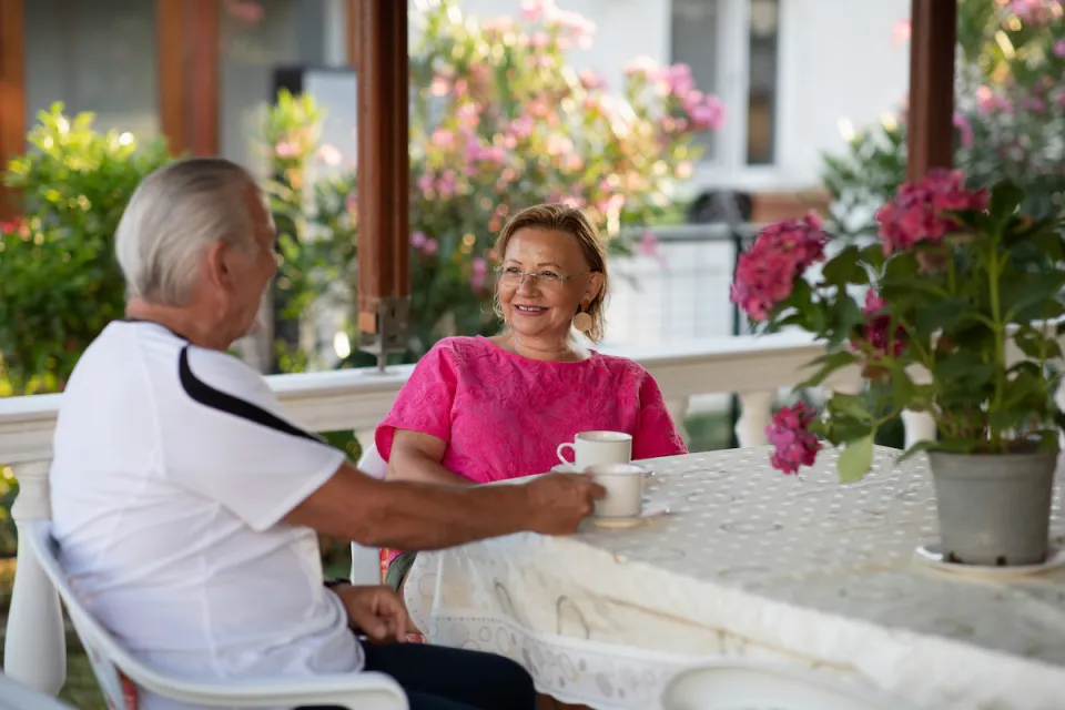 Elderly woman in pink shirt sitting at table on porch across from elderly man in white shirt. A pot of pink flowers sits on the table.